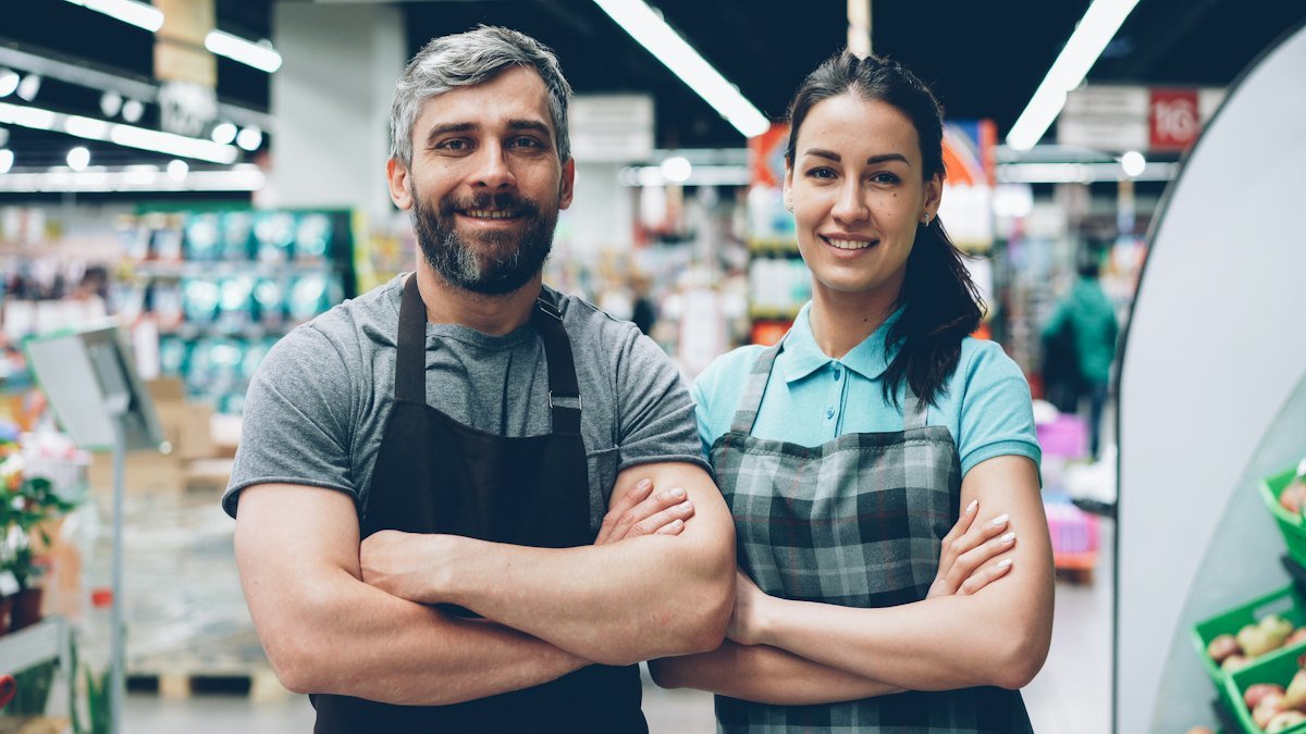 Dois donos de mercado sorrindo de braços cruzados entre as gôndolas