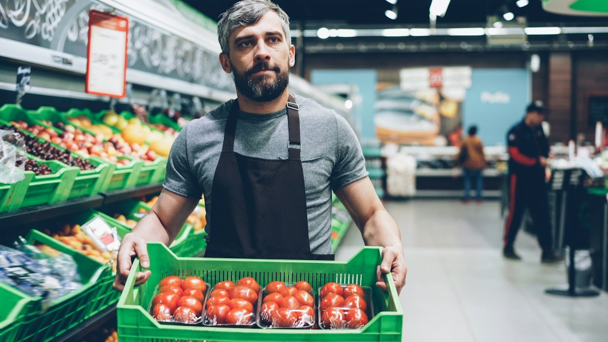 Funcionário de mercado segurando uma caixa de tomates no setor de hortifrúti